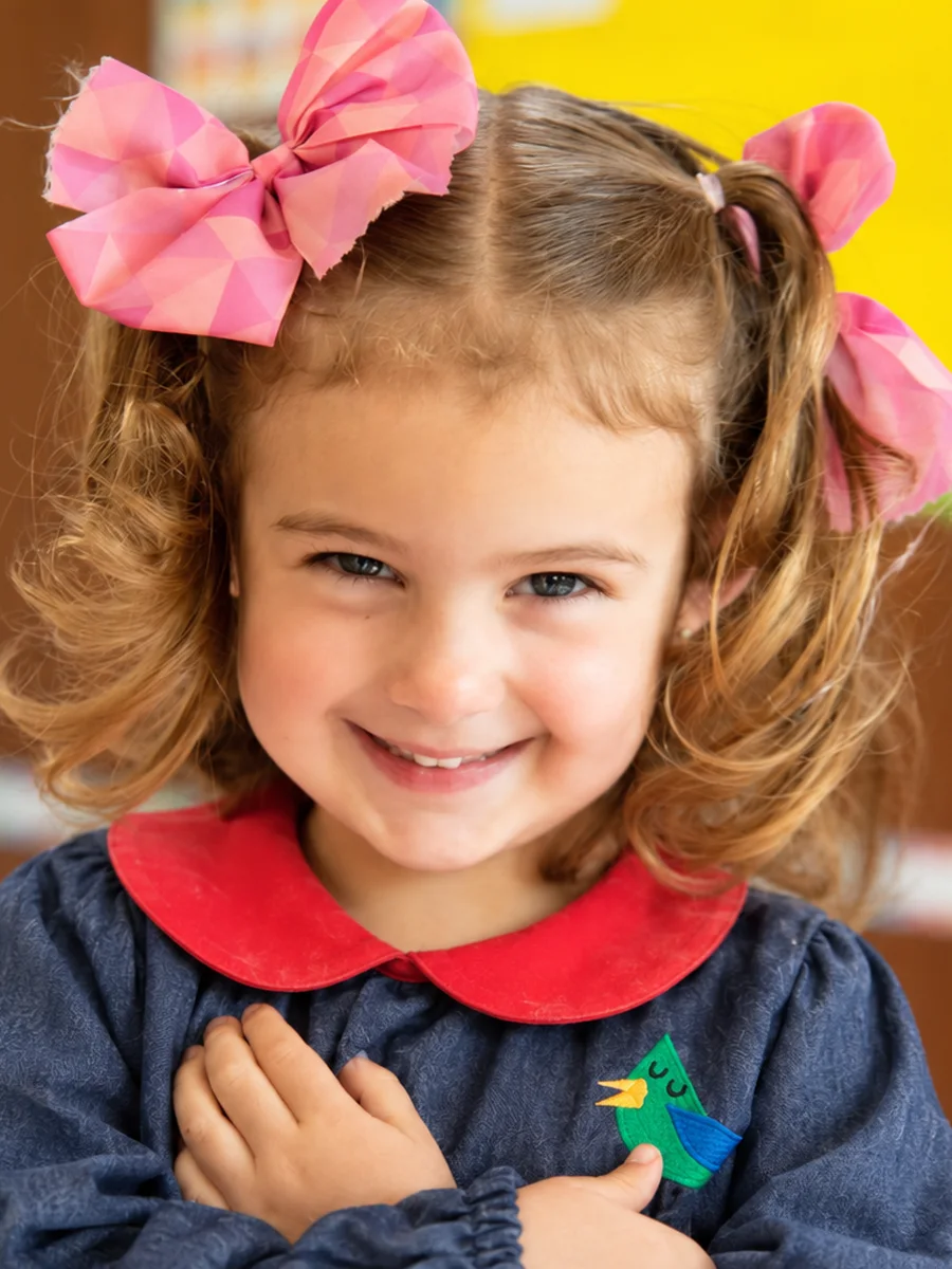 Niña de Piruli con moños rosas y su bata del uniforme, sonriendo con las manos en el pecho